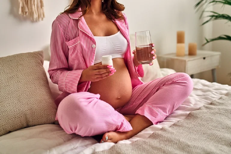 pregnant women sitting on her bed with a glass of water in one hand and a bottle of prenatal vitamins in the other waiting to take her daily dose