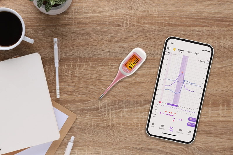 bbt thermometer on desk next to phone showing the premom app for online tracking